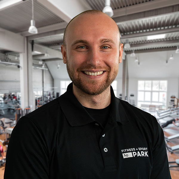 Lukas, Bewegungscoach von f+p Gesund Bewegen, im modern ausgestatteten Fitnessstudio im Allgäu, trägt schwarzes Polo mit PARK-Logo und lächelt freundlich in die Kamera.