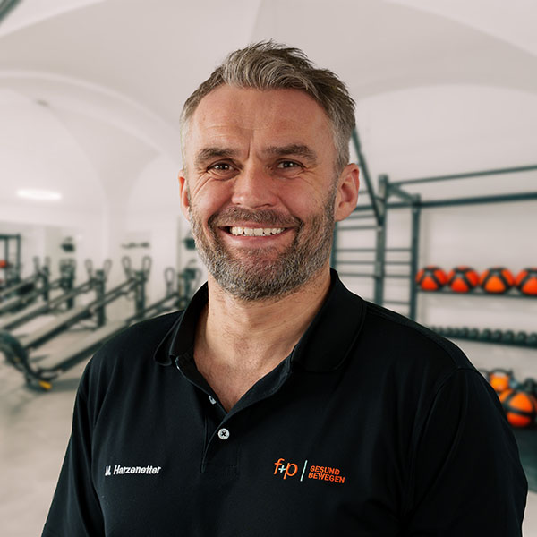 Martin von f+p Gesund Bewegen im Allgäu, trägt ein schwarzes Polo mit Logo, lächelt frontal in einem Fitnessstudio mit Trainingsgeräten im Hintergrund, Allgäu, Deutschland