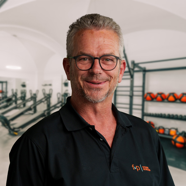 Marco, Facharzt für Chirurgie und Unfallchirurgie, trägt eine schwarze Polo-Hemd mit f+p Logo, graue Haare und Brille, steht vor einem Trainingsraum mit Geräten in Kempten