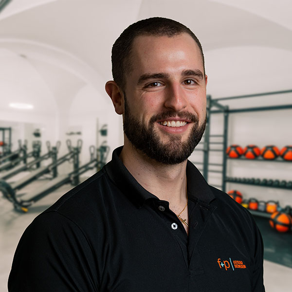 MaBu Manuel, ein freundlicher Mann mit dunklem Bart und kurzen Haaren, trägt ein schwarzes Polo-Shirt der Praxis f+p Gesund Bewegen und steht vor Trainingsgeräten im hellen Fitnessraum der Klinik im Allgäu.