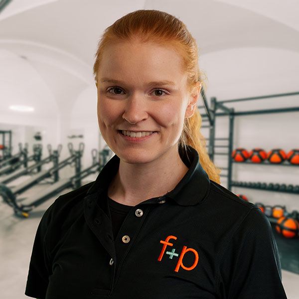 Magdalena von f+p Gesund Bewegen im Allgäu, Portrait im Trainingsraum mit orangem Logo f+p auf schwarzer Polo-Bluse, freundliches Lächeln, helles Haar