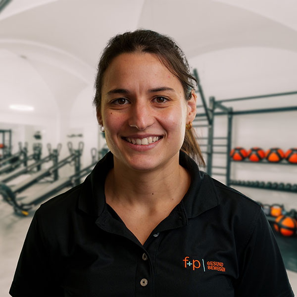 Martina, eine freundliche Physiotherapeutin von f+p Gesund Bewegen im Allgäu, trägt ein schwarzes Polo im Trainingsstudio, im Hintergrund stehen Trainingsgeräte und Rotationsbälle.