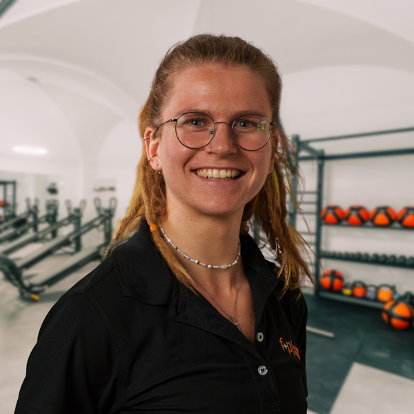Maike von f+p Gesund Bewegen mit blondem Haar und runder Brille, freundliches Lächeln, schwarze Bluse im Trainingsstudio mit Geräten und Hanteln im Hintergrund.