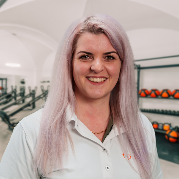 Melany, Mitarbeiterin von f+p Gesund Bewegen aus dem Allgäu, mit lavendelfarbenen Haaren und weißem Hemd vor einem Fitnessraum.
