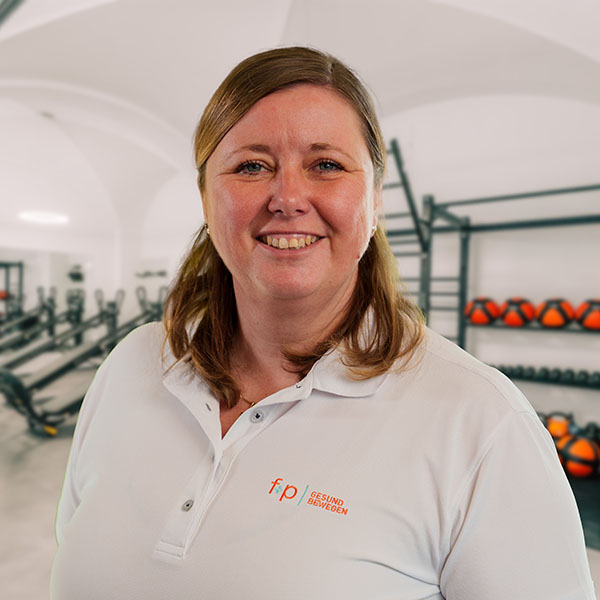 Miriam von f+p Gesund Bewegen im Allgäu, lächelndes Portrait in weißem Polohemd vor Fitness-Equipment, Logo fp Gesund Bewegen sichtbar auf der Brust, ruhige klare Bildstimmung