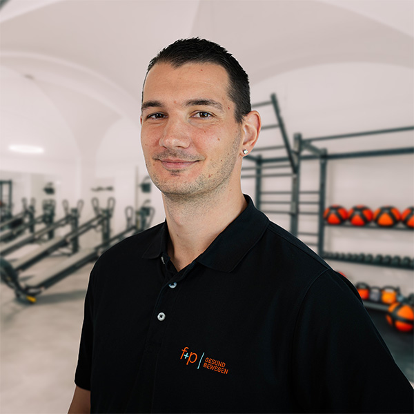 Mann mit kurzen Haaren und Ohrringen, trägt ein schwarzes Poloshirt mit Logo vor einer Fitnessumgebung.