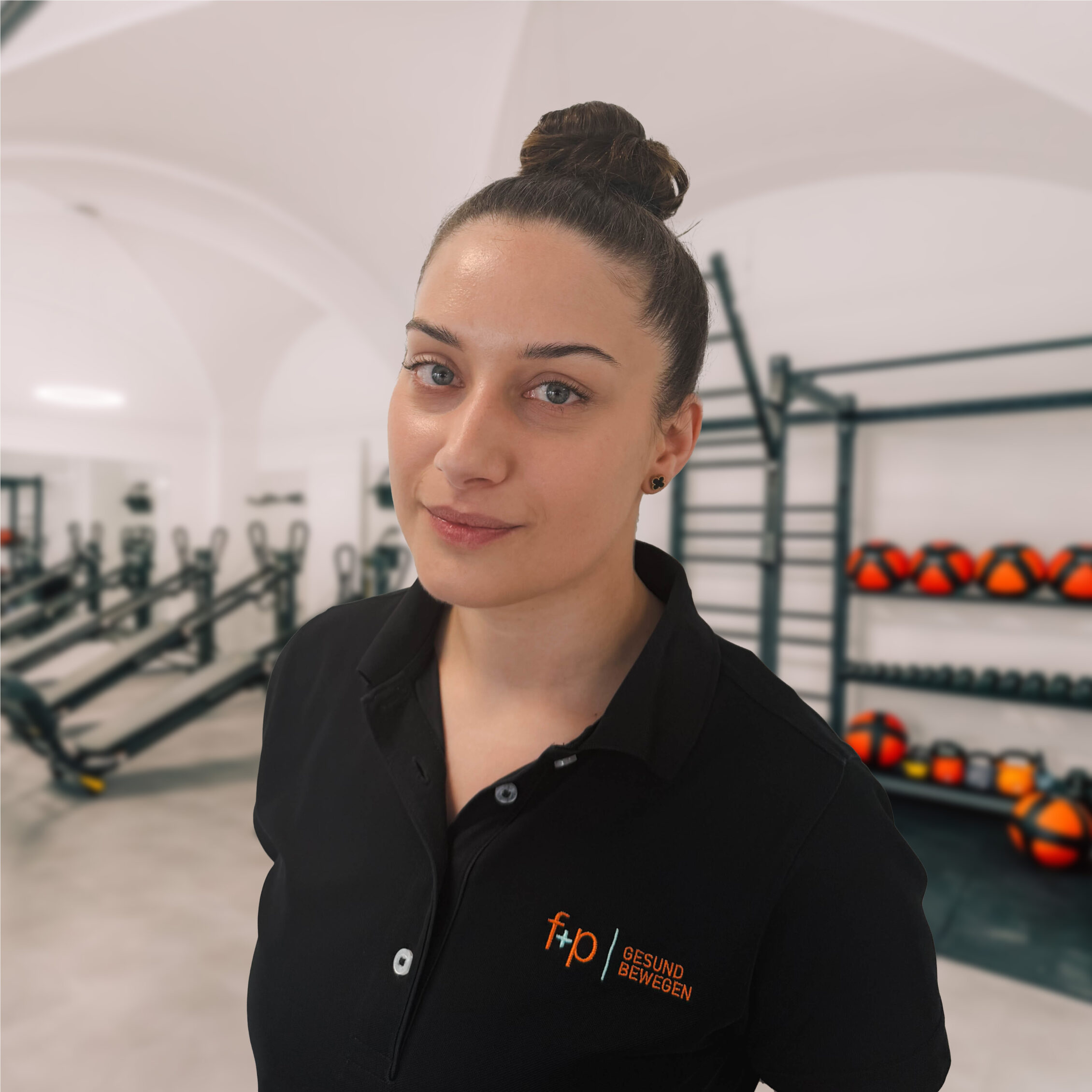 Pamela von f+p Gesund Bewegen mit freundlichem Blick in schwarzer Kleidung, im Hintergrund Trainings- und Reha-Bereich mit Geräten und Hanteln.