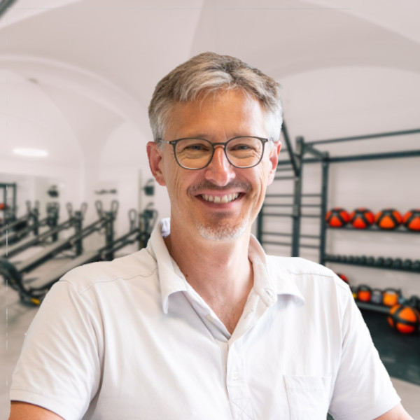 Lächelnder Mann mit Brille in einem Fitnessstudio mit Fitnessgeräten im Hintergrund.