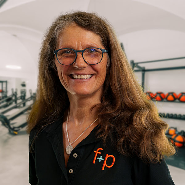 Regina, eine Frau mit braunen Haaren und blauer Brille, lächelt im Fitnessstudio von f+p Gesund Bewegen im Allgäu; Hintergrund Trainingsbereich mit Geräten erkennbar.