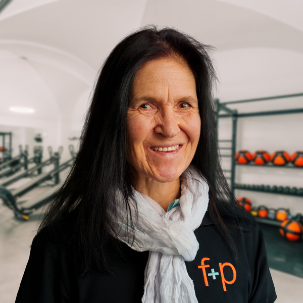 Sabina mit schwarzen Haaren und weißem Schal lächelt in einem Trainingsraum, im Hintergrund Trainingsgeräte und rote Medizinbälle sichtbar