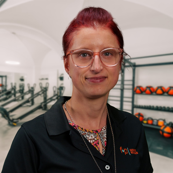 Saskia mit roten Haaren und leichter Brille, Portrait im Trainingsraum von f+p Gesund Bewegen im Allgäu, im Hintergrund Fitnessgeräte und Rogor-Trainingsbanken.