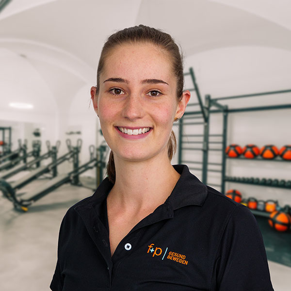 Sabrina von f+p Gesund Bewegen im Allgäu lächelt in einem Fitnessstudio, trägt ein schwarzes Polo-Shirt mit Logo, im Hintergrund Trainingsgeräte