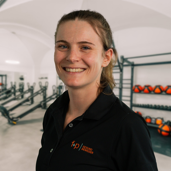 Sabrina in schwarzer Polo-Blusenkleidung von f+p Gesund Bewegen im Allgäu, lächelnd im Trainingsraum mit Trainingsgeräten und red/orange Hantelscheiben im Hintergrund, Allgäu