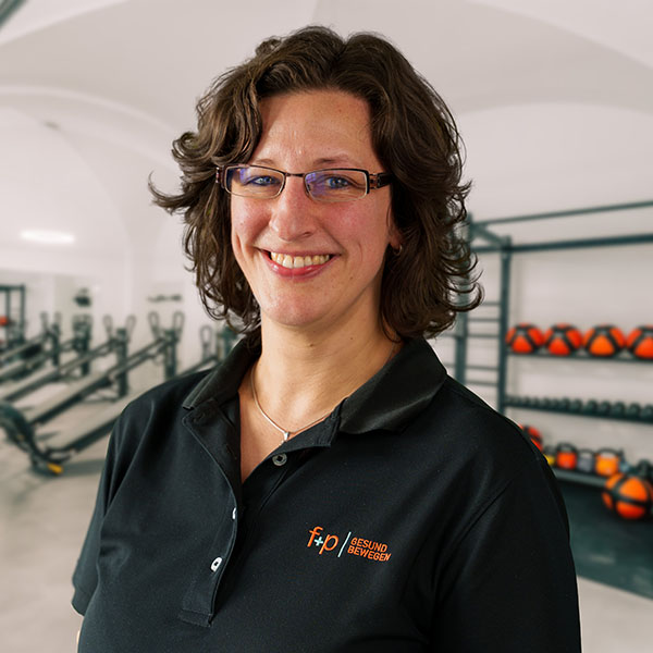 Sarah, Mitarbeiterin bei f+p Gesund Bewegen im Allgäu, trägt schwarzen Polo mit Logo, trägt Brille und lächelt in einem Therapieraum mit Fitnessgeräten im Hintergrund.