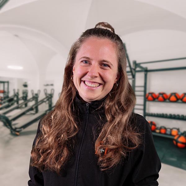 Selina in einer hellen Physiotherapie-Praxis, braune lange Haare, schwarze Jacke, lächelnd, im Hintergrund Trainingsgeräte wie Kettlebells und Therapieräder