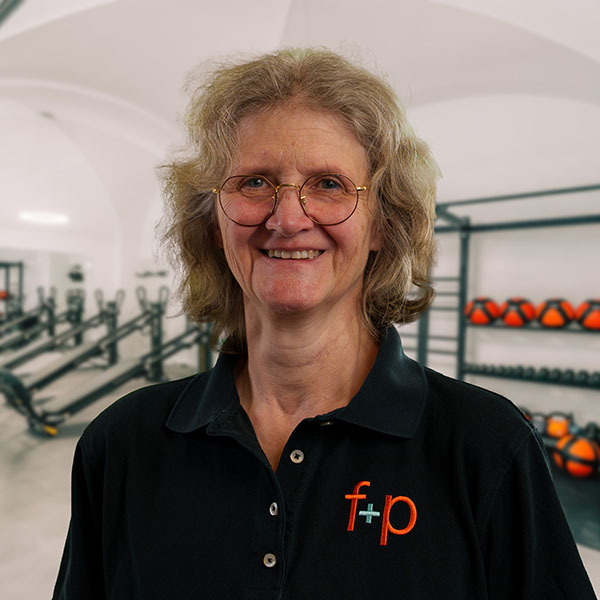 Stefanie, Physiotherapeutin bei f+p Gesund Bewegen im Allgäu, lächelt freundlich im Trainingsraum; porträtierte Person mit Brille, heller Haarfarbe und dunklem Poloshirt mit dem f+p Logo