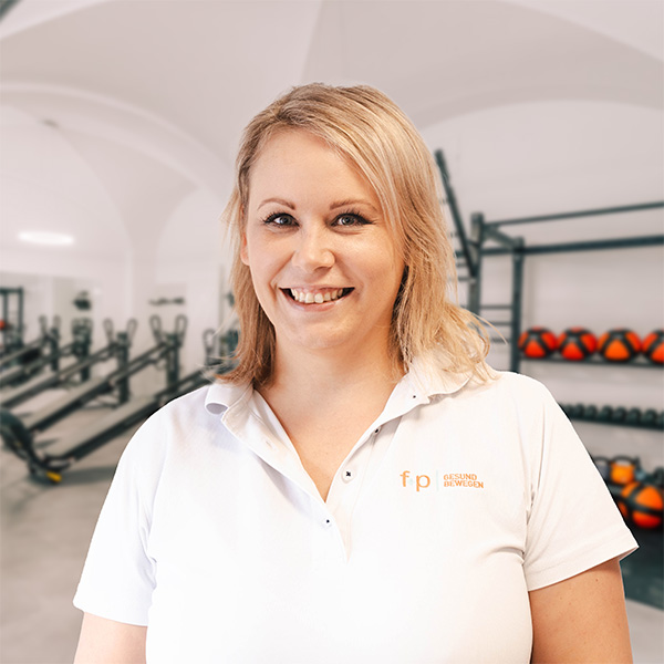 Frau mit blonden Haaren in einem weißen Poloshirt lächelt vor einem Fitnessstudio-Hintergrund.