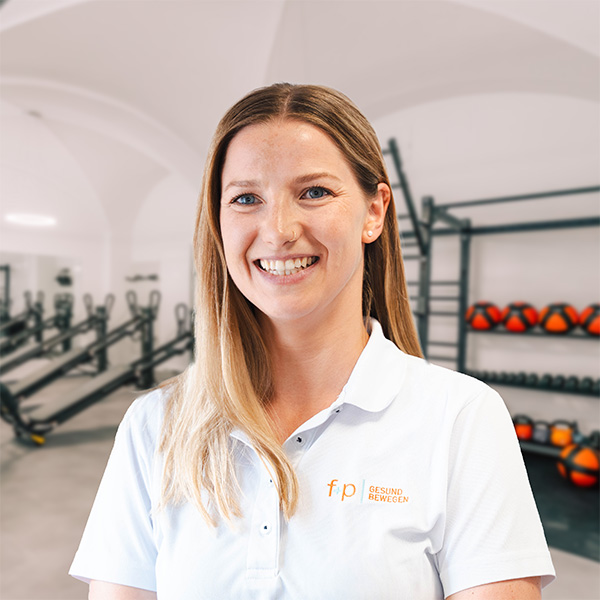 Tanja, eine Frau mit hellem Haar, trägt ein weißes Polohemd mit dem Logo f+p Gesund Bewegen, im Hintergrund eine gym-ähnliche Einrichtung mit Trainingsgeräten.