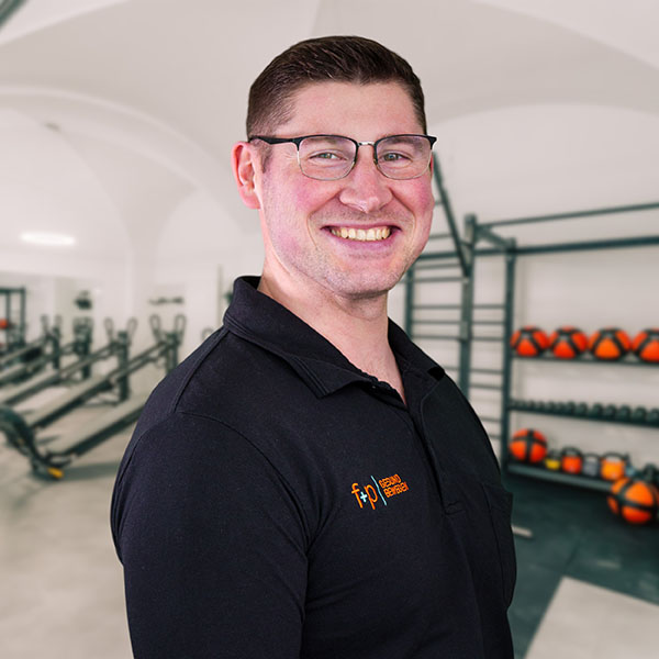 Thomas, ein lächelnder Mann mit Brille in einem schwarzen Polo der Marke f+p Gesund Bewegen, steht im Trainingsraum im Allgäu mit Fitnessgeräten im Hintergrund.