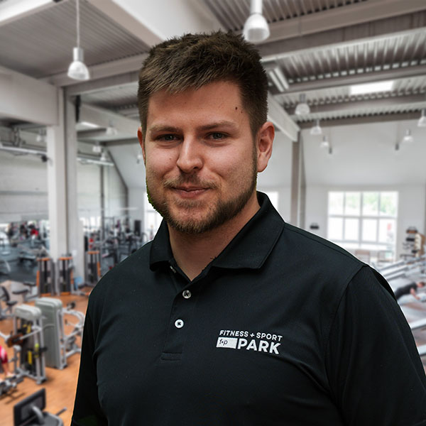 Toni, ein braunhaariger Mann mit kurzem Bart, trägt ein schwarzes Polo mit dem Logo Fitness & Sport Park und steht in einem hellen Fitnessstudio mit Gerätschaften im Hintergrund.