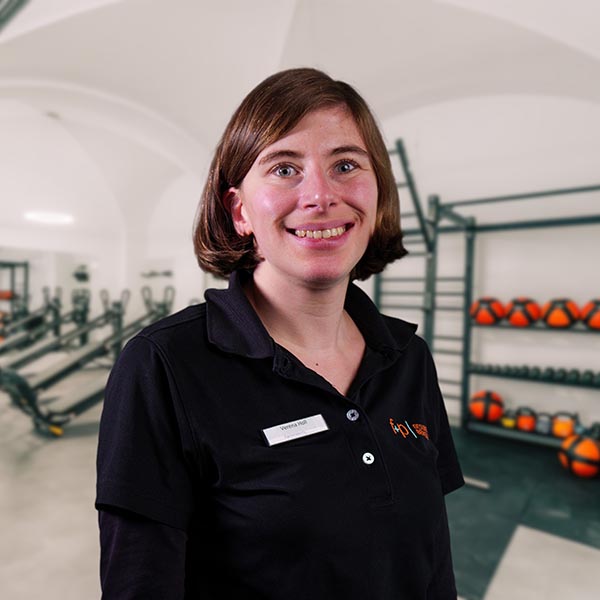 VeHo Verena, Ergotherapeutin bei f+p Gesund Bewegen im Allgäu, lächelt in einem schwarzen Polo – im Hintergrund Trainingsgeräte wie Hanteln und ein Klettergerüst sind sichtbar.