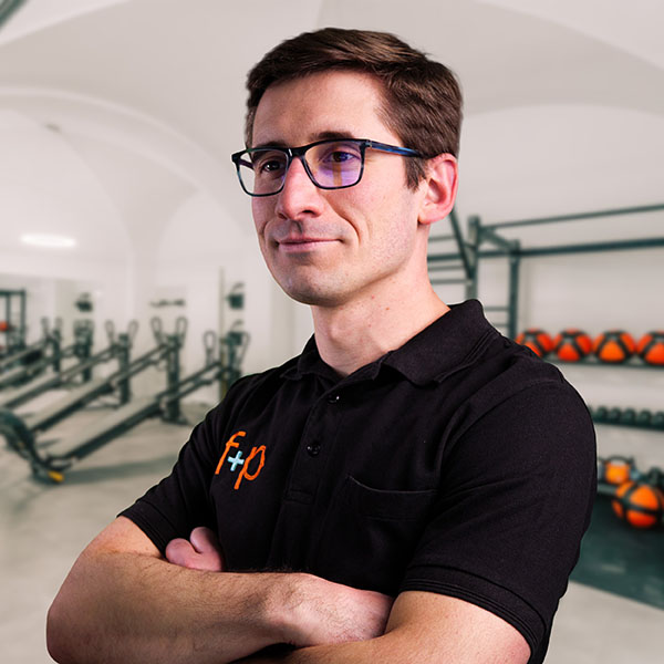 Vladimir, ein Mann mit dunklem Polohemd und Brille, steht mit verschränkten Armen in einem Fitnessstudio der f+p Gesund Bewegen im Allgäu, Hintergrund mit Trainingsgeräten.
