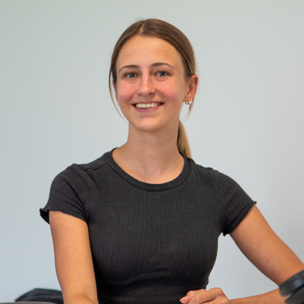 elisable Mit Elisa von f+p Gesund Bewegen steht eine freundliche Mitarbeiterin mit Ponytail und schwarzem Shirt vor hellem Hintergrund im Allgäu.