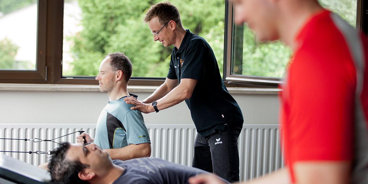Ein Physiotherapeut führt eine Behandlung bei einem Patienten durch, während ein anderer Patient im Hintergrund trainiert.