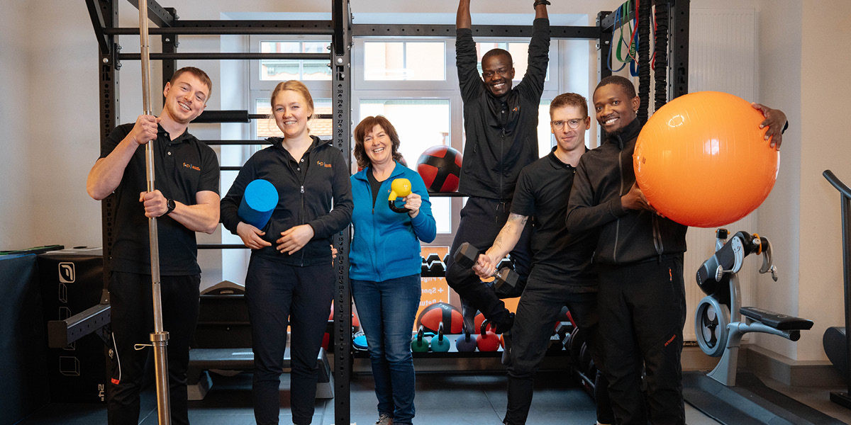 Das Team von f+p Gesund Bewegen posiert mit Fitnessgeräten in einem Trainingsraum.
