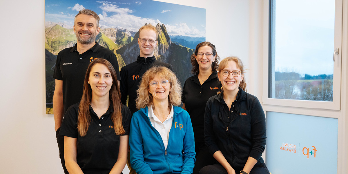 Sechs Teammitglieder von f+p Gesund Bewegen posieren in einem Raum mit Berglandschaft im Hintergrund.