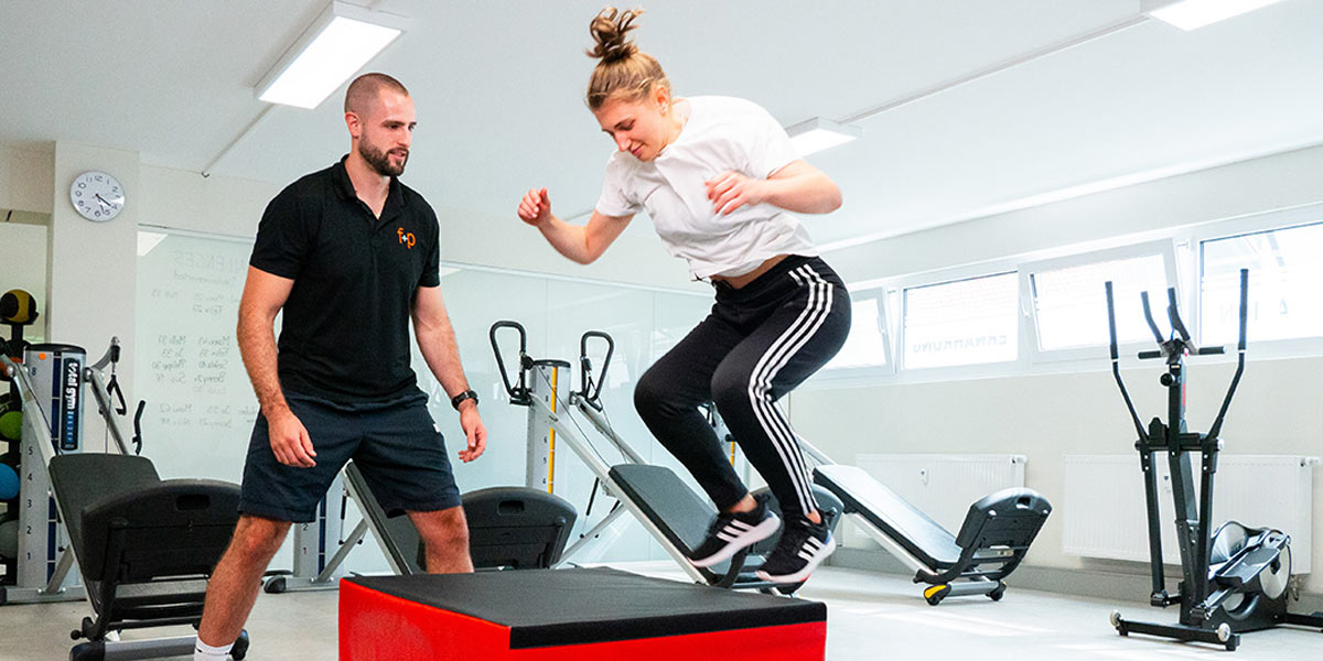 Eine Person springt auf einen roten Trainingsblock, während ein Trainer daneben steht und beobachtet.