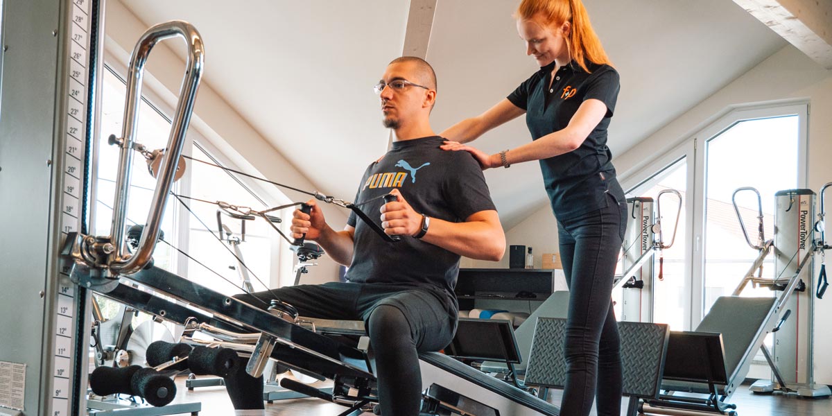 Mann im Fitnessstudio, der an einem Trainingsgerät sitzt, während ein Trainer ihn unterstützt.
