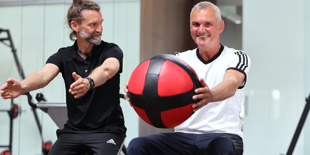 Ein Trainer und ein älterer Mann im Fitnessstudio, der einen Medizinball hält und Übungen macht.