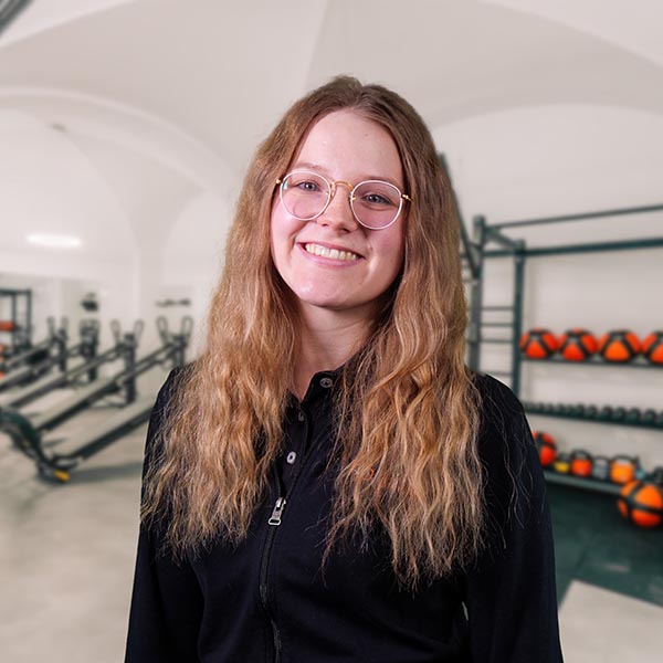 Junge Frau mit langen, lockigen Haaren und Brille in einem Fitnessstudio.
