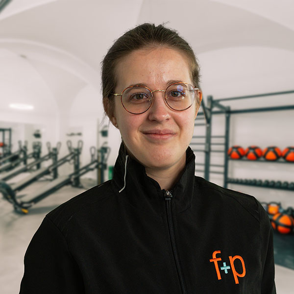 Frau mit Brille und schwarzer Jacke steht vor einer Sporteinrichtung mit Fitnessgeräten.