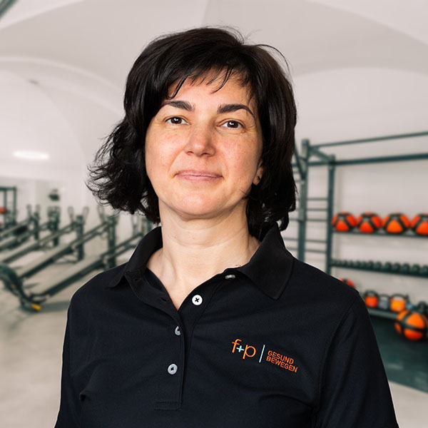 Andreea, Fachärztin für Physikalische und Rehabilitative Medizin bei f+p Gesund Bewegen, steht in Trainingsraum mit schwarzem Polo, Allgäu-Logo sichtbar, kompetent wirkend
