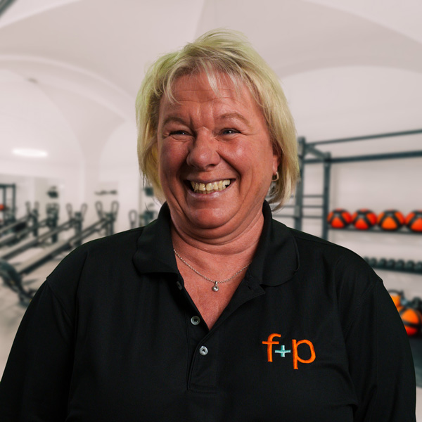 Angelika Schmid Lächelnde Frau in schwarzer Sportbekleidung mit Logo, vor einer Fitnessumgebung.