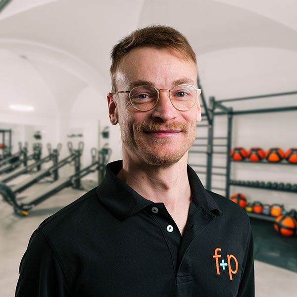 Bastian, Mitarbeiter von f+p Gesund Bewegen, trägt eine schwarze Polo-Hemdjacke und Brille. Hintergrund mit Trainingsgeräten im Allgäu-Gym.