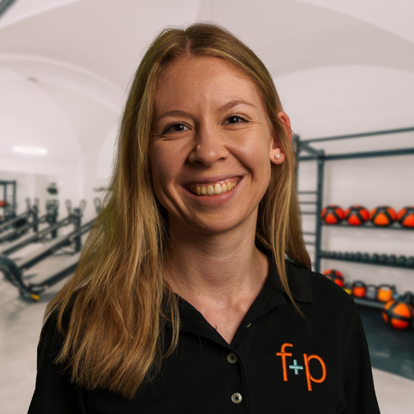Carina Gerber Frau mit langen blonden Haaren in einem schwarzen Hemd mit Logo, lächelnd vor einem Fitnessstudio-Hintergrund.