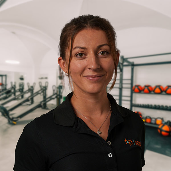 Frau mit braunen Haaren in einem Fitnessstudio, trägt ein schwarzes Polohemd.