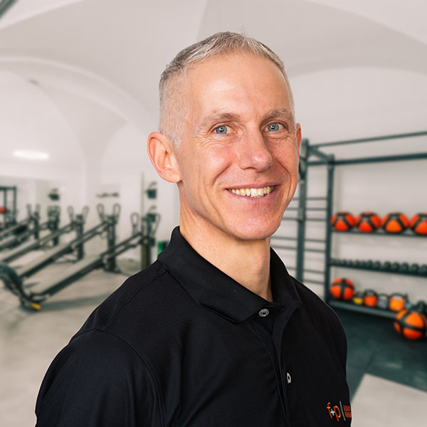 Christian Dörr Ein Mann mit kurzem, grauem Haar und schwarzem Polohemd lächelt in einem Fitnessstudio mit Trainingsgeräten im Hintergrund.