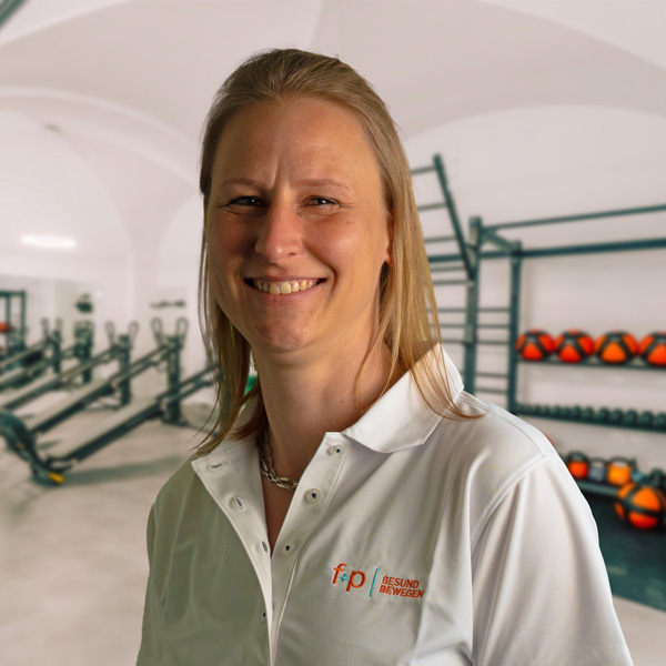 Frau mit blonden Haaren in einem weißen Poloshirt steht in einem Fitnessstudio mit Trainingsgeräten im Hintergrund.