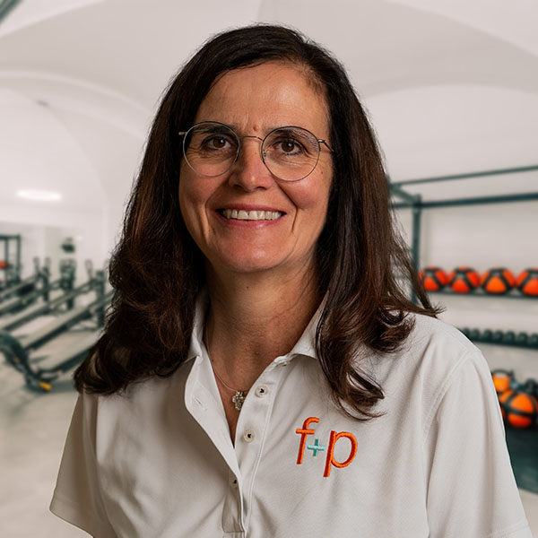 Frau mit Brille und weißem Polohemd steht in einem Fitnessstudio mit Fitnessgeräten im Hintergrund.