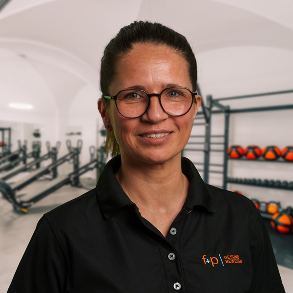 Claudia-Re, Mitarbeiterin von f+p Gesund Bewegen im Allgäu, trägt eine schwarze Polo-Bluse, Brille und steht in einem Trainingsraum mit Geräten und Hanteln.