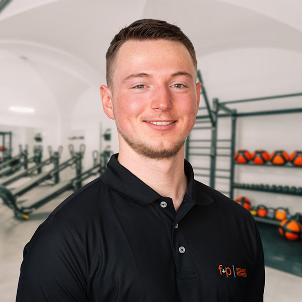 Daniel, Mitarbeiter bei f+p Gesund Bewegen im Allgäu, lächelt freundlich und trägt ein schwarzes Polo mit dem Logo von f+p; im Hintergrund befindet sich ein modern ausgestattetes Fitness- und Therapiezentrum.