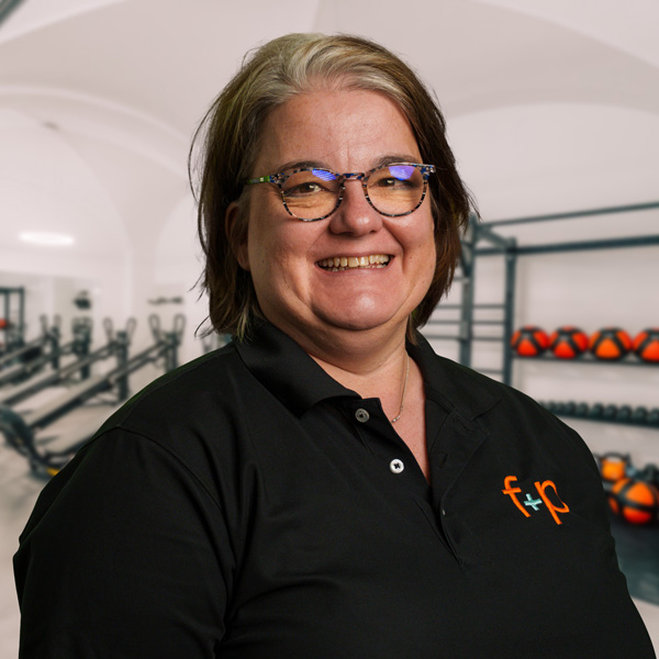 Daniela Le von f+p Gesund Bewegen, lächelnd, trägt schwarzes Polo mit orangeem Logo, Brille, Trainingsraum im Hintergrund mit Geräten