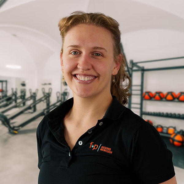 Frau mit lockigem Haar in schwarzer Sportbekleidung steht in einem Fitnessraum mit Geräten.