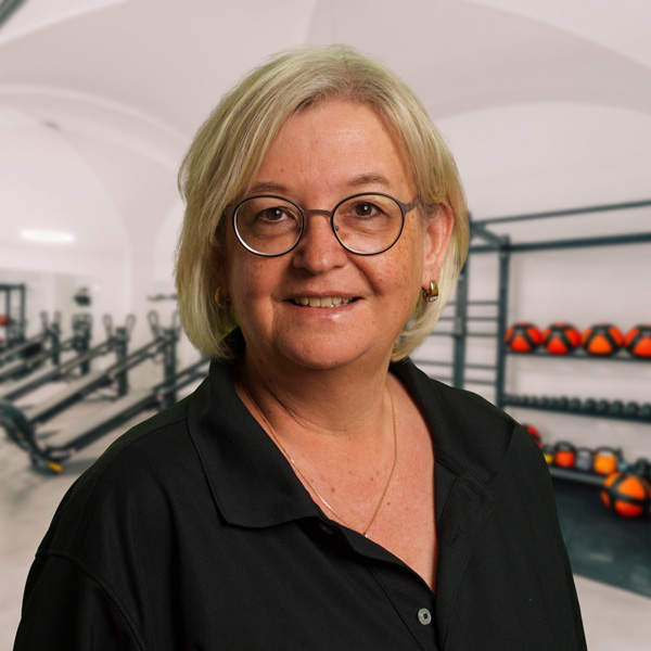 Elke, blondes Haar, runde Brille, schwarzes Oberteil, steht vor Trainingsgeräten in einem Fitnessraum mit Hanteln sichtbar im Hintergrund.