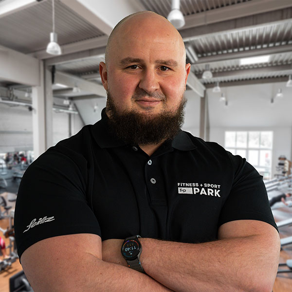 Mann mit Bart trägt ein schwarzes Fitness-T-Shirt und steht in einem Fitnessstudio mit Geräten im Hintergrund.
