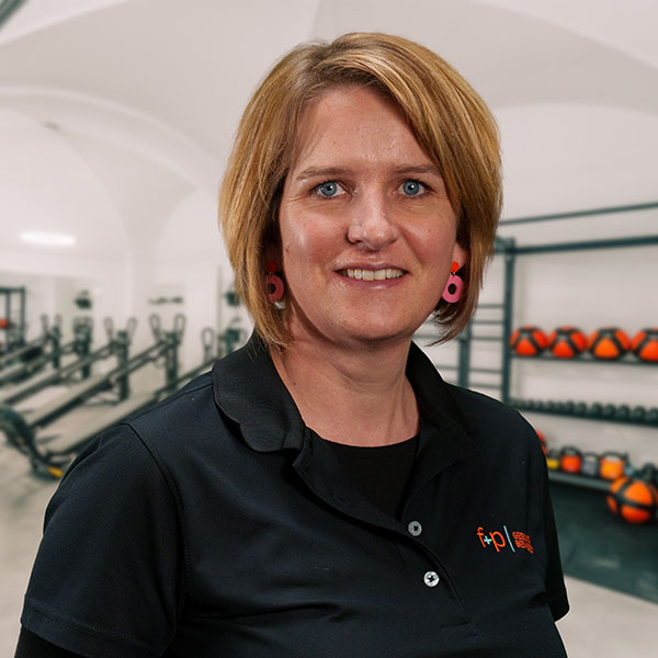 Frau mit blonden Haaren und schwarzem Sporthemd, lächelt in einem Fitnessstudio.